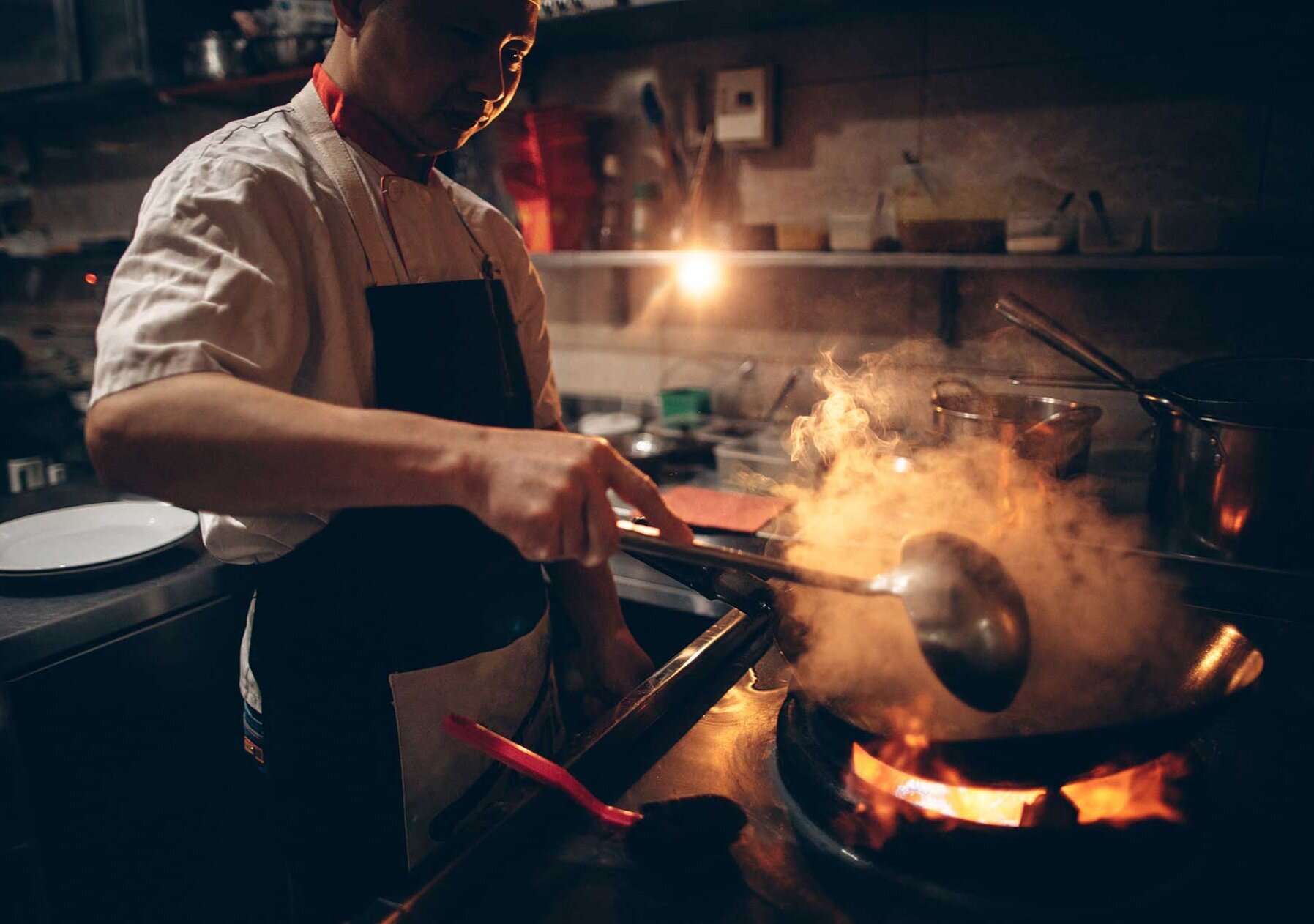 Chef stands in a dark kitchen in front of a steaming pan Chef stands in a dark kitchen in front of a steaming pan